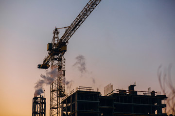 Crane and building construction site with pipe with smoke on background of sunset sky. Industrial landscape with silhouettes of cranes over sunset. Pollution from factory or power plant