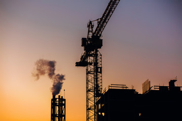 Crane and building construction site with pipe with smoke on background of sunset sky. Industrial landscape with silhouettes of cranes over evening sunlight. city Environmental problem