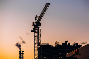 Fototapeta na wymiar Crane and building construction site with pipe with smoke on background of sunset sky. Industrial landscape with silhouettes of cranes over evening sunlight. city Environmental problem