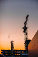 Crane and building construction site with pipe with smoke on background of sunset sky. Industrial landscape with silhouettes of cranes over evening sunlight. city Environmental problem