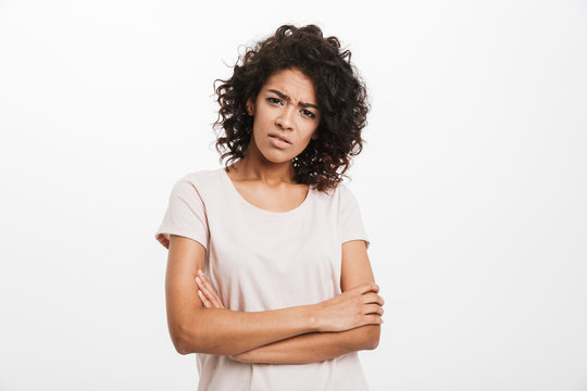 Closeup photo of young afro american woman 20s wearing casual t-shirt looking on camera with frown keeping arms folded, isolated over white background