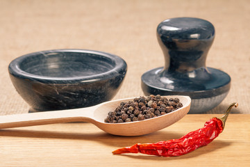 Wooden spoon with black peppercorn, dried red pepper on cutting board and stone mortar for grinding pepper