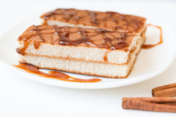Biscuit cake with caramel syrup on white plate with cinnamon sticks on wooden background, close-up