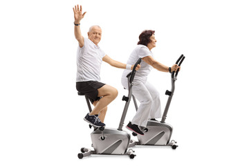 Elderly man and an elderly woman riding exercise bikes with the man waving