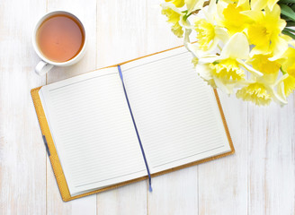 Top view Open notebook with blank pages Cup of Tea and Yellow Daffodils on Light Wooden Background. Still life, business, office supplies or education concept.