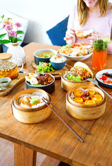 A large, wooden table. A young, beautiful girl in pink is sitting at the table. On the table there are many different dishes of French and Chinese cuisine: salads, dumpling, meat stew and many other