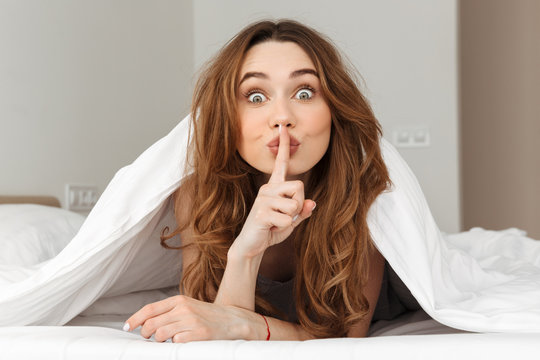 Photo of young brunette woman lying in bed under blanket at home, and gesturing finger on mouth to keep silence