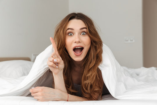 Photo of excited woman 20s lying in bed under white blanket in bedroom, and having idea putting index finger up