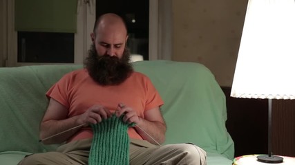A charismatic man with a beard knits a green scarf while sitting in a room on the couch. Bearded man trying to knit