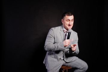 Handsome man in gray suit with microphone against black background on studio sitting on chair. Toastmaster and showman.