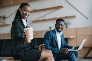 selective focus of smiling african american businessman with laptop and colleague showing coffee to go near by in cafe