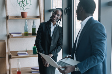 smiling african american businessman with laptop and colleague near by in cafe