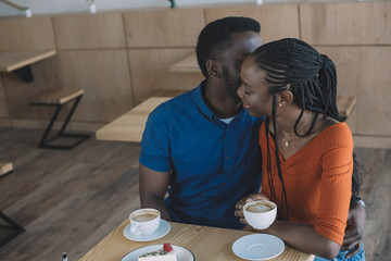 tender african american couple sitting at table with cups of coffee and dessert in cafe