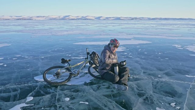The man is resting sitting on the wheel of a bicycle on ice. He drinks tea from a thermos bottle. The cyclist is dressed in a gray down jacket, backpack and helmet. Ice of frozen Lake Baikal. The