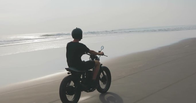 Handsome man biker driving his black motorcycle cafe racer on the beach along the ocean during sunset. 4k video shooting by handheld gimbal