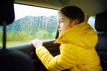 Adorable girl sitting in a car and looking outside on rainy autumn day. Child entertaining herserf on a road trip.