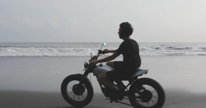 Handsome man biker driving his motorcycle cafe racer on the beach along the ocean during sunset. 4k video shooting by handheld gimbal