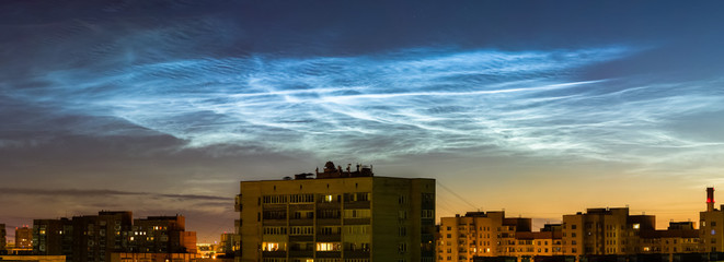 Wide panoramic night sky view of beautiful noctilucent clouds over the city with a cityscape roofs at foreground, rare phenomena which happens only on summer nights. cityline with awesome cloudy sky