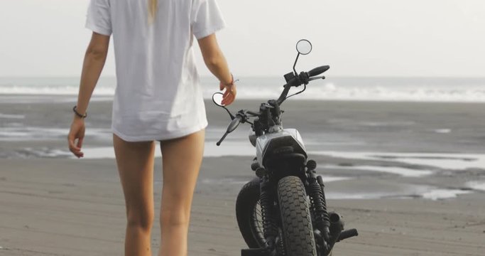 Pretty girl wearing white clothes sits down on vintage custom motorcycle cafe racer traveling and having fun. Young female biker woman posing on beach bali. 4k video shooting by handheld gimbal