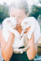 Beautiful young woman holds two white cats in her arms and kisses them in the sun light