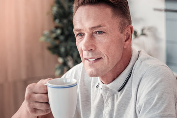 Positive thoughts. Attractive male person keeping smile on his face and holding cup in right hand while looking forward