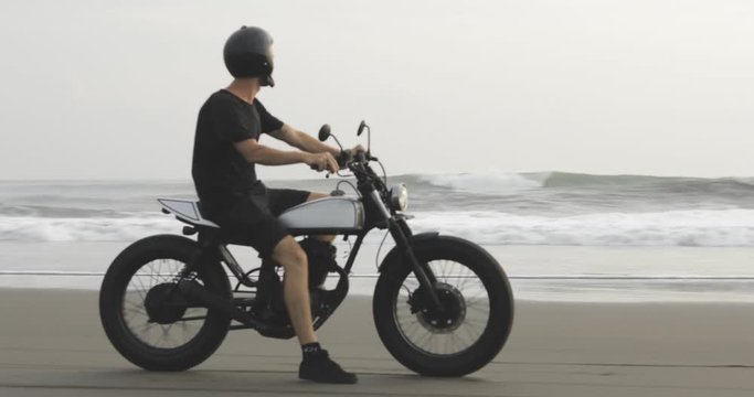 Handsome man biker driving his motorcycle caferacer on beach along ocean during sunset. 4k video shooting by handheld gimbal