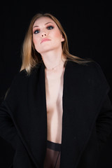 Sexy lady with glamour makeup and long blonde hair in nylons and long coat undressing, in studio. Elegant woman wearing black sensual lingerie. Erotic noir concept. 