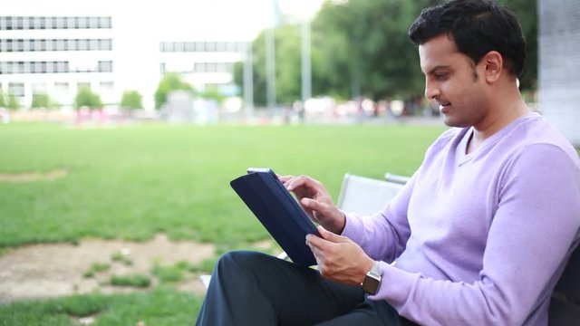 Closeup portrait, happy handsome young guy in purple sweater and black pants, sitting on bench playing with touch-screen tablet pc, isolated outdoors outside background. Positive facial expressions