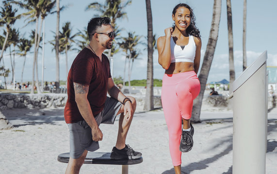 Laughing African-American girl in sportswear doing exercise under control of cheerful sportive man training on beach.