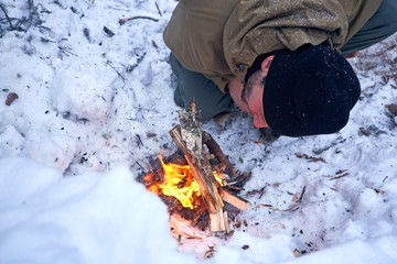 Young man in the winter forest bends over a small fire and and blowing on embers