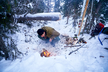 Young man in the winter forest bends over a small fire and and blowing on embers