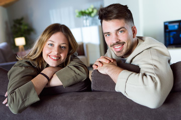 Beautiful young smiling couple looking at camera and sitting on sofa at home.