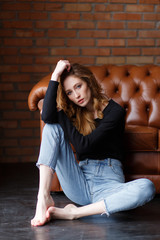 Fototapeta na wymiar Young woman in jeans and pullover sitting on floor near brown leather sofa