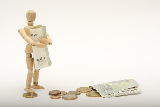 A wooden person stands and holds money, on the ground lying coins and euro banknotes (money) (person looks at the viewer)