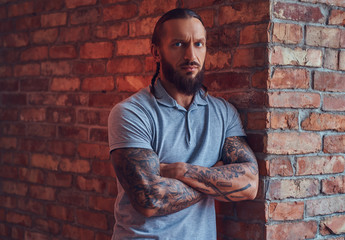Obraz premium A handsome tattoed male with a stylish haircut and beard, in a gray t-shirt, standing leaning against a brick wall in a room with a loft interior.