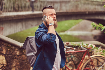 Portrait of a bearded male with a haircut dressed in casual clothes with a backpack, standing in a park, talking by phone.