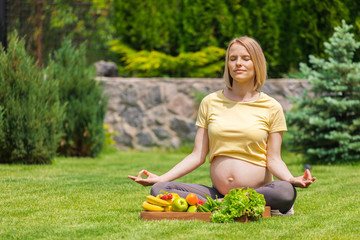 A pregnant woman sits on the grass with a tray of fresh fruits and vegetables, meditates and relaxes in the fresh air. The concept of a healthy lifestyle. Caring for yourself and future child