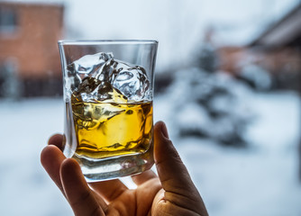 Glass of whiskey with ice cubes  in man's hand. Winter nature at the background.