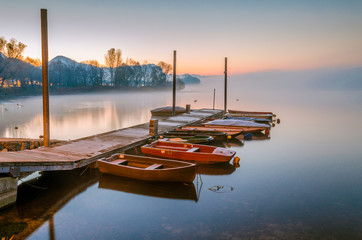 Boats moored in the small pier on the Adda river in Calolziocorte near Lecco. The sun is about to rise on a foggy morning - obrazy, fototapety, plakaty