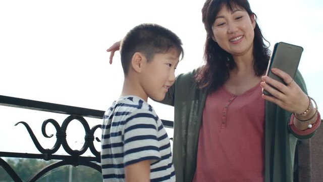 Asian woman and little boy smiling and posing at camera of smartphone while taking selfie together outdoor at sunny day