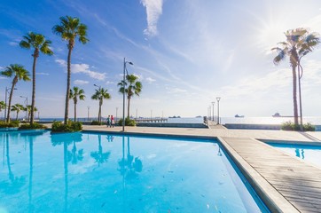 Molos Promenade and skyline of the coast in Limassol city in Cyprus at sunrise. View of the boardwalk pier path landmark with palm trees, pools of water, the Mediterranean sea and people walking.