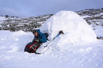 hiker pours himself a hot drink from a thermos, sitting in a snowy house igloo against a background...