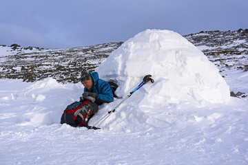 hiker pours himself a tea from a thermos, sitting in a snowy hut igloo against a background of a winter mountain landscape..