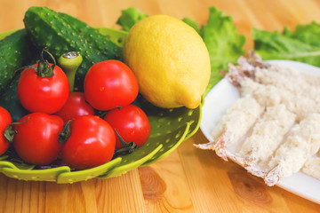 Raw shrimp in batter ready for roasting next to a basket of cucumber lemon cucumbers and tomatoes against the background of lettuce leaves on a wooden table