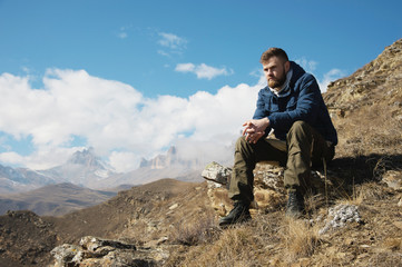 A portrait of a stylish bearded hipster sitting on a rock against the backdrop of epic rocks and contemplating into the distance thinking about life. The concept of tranquility and solitude with
