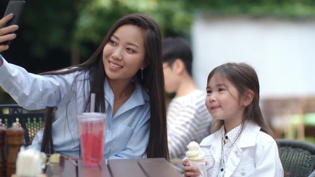 Tracking shot of young pretty Asian woman taking selfie with cute little girl posing with ice cream at smartphone camera in outdoor cafe