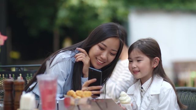 Tracking shot of cute Asian little girl sitting with ice cream at cafe table outdoors and posing at smartphone camera while woman taking picture of her