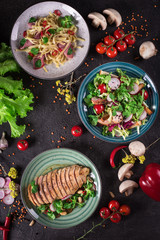 Delicious dinner. Italian pasta, fresh vegetable salad and roasted chicken breast on the black background. Healthy food. Close up. Top view