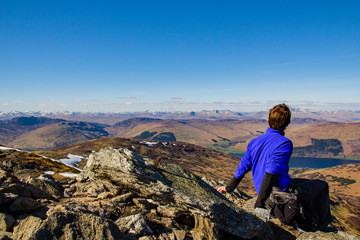 A male hiker sits upright and rests and looks in to the distance on a clear bright sunny day at the...
