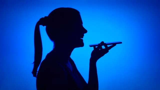 Silhouette of young woman using mobile on blue background. Female's face in profile chatting with friend on cellphone. Black contur shadow of teenager's half-face talking